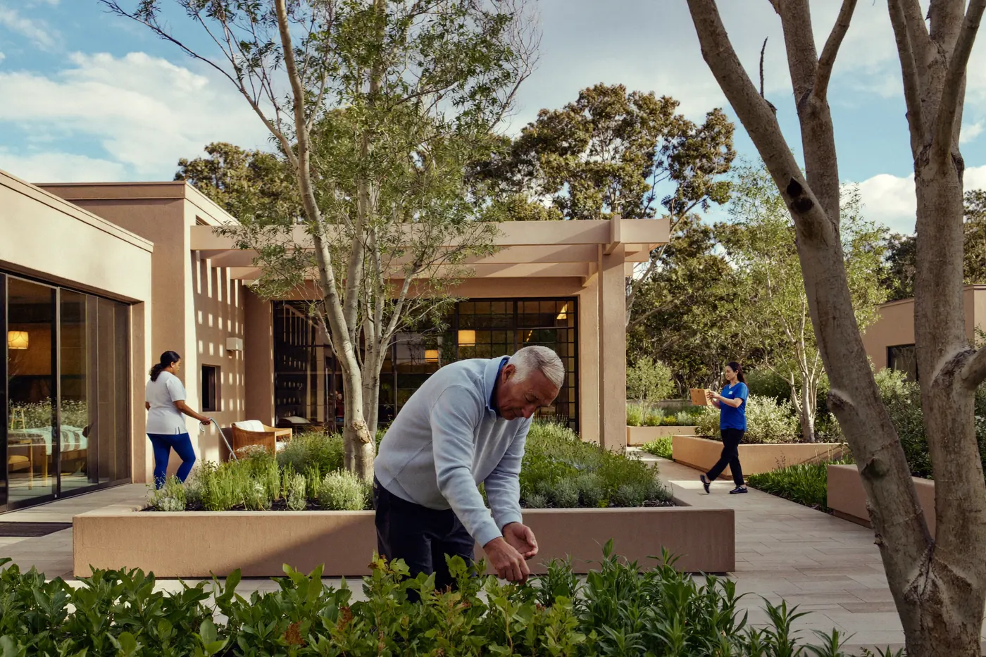 An older man tends herbs in a secured memory care courtyard garden with staff nearby